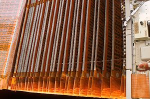 Close up of folded P6 solar array, individual cells sandwiched into orange film, short conducting connections can be seen between cells, with straight conductors called Flexible Flat Conductor (FFC) Cable or Flexible Film Connectors similar to those used in domestic electronics and flat panel LCD connections. 38 white piano hinges are apparent, only those closest to the camera are visible. The ends of the folded film array are attatched by small springs, 17 of a probable 40 are visible. Mounting has label giving foot pound tensioning instructions for latch tension and delatching detension, with manual, automatic labels. Solar 'blanket' contains 16,400 cells