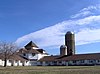 Two wings of a white barn with two silos under a blue sky with cirrus clouds