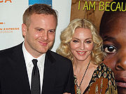 A blond middle-aged woman and a middle-aged man stand beside each other, smiling towards the camera. The woman wears a brown kaftan hwile the man is in black suit.