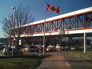 An overhead walkway is above several rows of cars waiting to cross the border