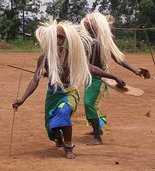 Foto yang menggambarkan dua penari pria dengan rambut palsu dari jerami, garmen leher, tombak dan tongkat.