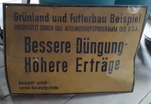 German sign indicating "agriculture counseling supported by the overseas aid program of the U.S.A." Schild Bessere Dungung des Amerikanischen Hilfsprogramms.png
