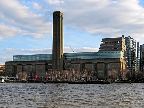 A large oblong brick building with square chimney stack in centre of front face. It stands on the far side of the River Thames, with a curving white foot bridge on the left.