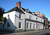 Photograph of a white-painted house; the main face has two storeys and attics, with an ornate doorway. The end section is two storeys with a large red-brick chimney.