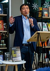 Yang is holding a microphone while gesturing and making a speech. His book, The War on Normal People, is displayed on a table in front of him.
