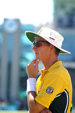 A man in a yellow cricket uniform and white sun hat stands with a his hand on his chin whilst fielding in a cricket match