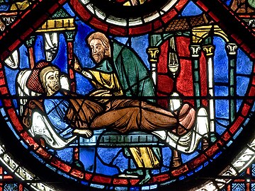 Mozaikové okno zobrazující zraněného muže vleže, který je ošetřen jiným mužem.
