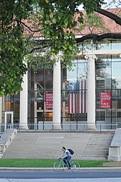 A photo of the John Glenn College of Public Affairs, with an American flag hanging inside and a cyclist riding past the stone steps