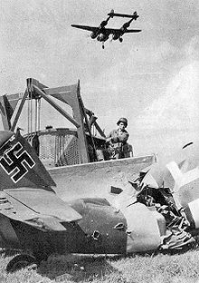 A USAAF Engineer clearing out the wreckage of a destroyed Luftwaffe Messerschmitt Bf 109 aircraft at an ALG, with a Lockheed P-38 Lightning flying overhead on landing approach IXengeering-enenemyairfield.jpg