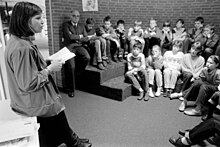 A black-and-white photo of a white woman with dark hair reading a book to an audience of children and adults