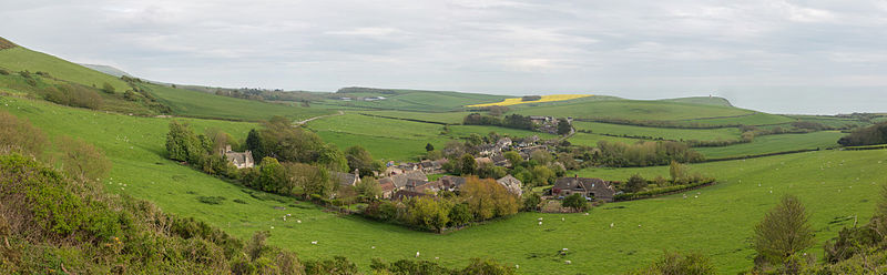 A panoramic view of the village from the north, looking towards the English Channel coast, which forms the horizon. The slopes of Smedmore Hill are to the left, and the Clavell Tower can just be seen on the step-shaped hill above and to the right of the village. The small parish church is on the left edge of the main cluster of buildings. Kimmeridge civil parish covers the village and the land beyond and to the left; the land in the foreground, between the camera and village, is part of neighbouring Steeple parish.