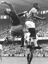 Photographie en noir et blanc de deux joueurs de football, un attaquant et un gardien de but, se disputant le ballon dans les airs.
