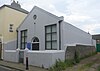 Three-quarter view of a small stuccoed building with a pediment. The longer side is a blank wall; a flint boundary wall runs alongside it. The façade has two simple rectangular windows flanking a round-arched entrance, and a small round window below the pediment.