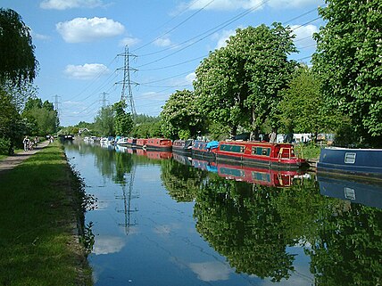 A wide river on a sunny day: on the left, cyclists and pedestrians on the towpath, on the right, canal boats