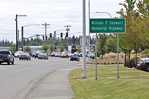 A wide suburban road with several parking lots. At the right of center is a sign reading "William P. Stewart Memorial Highway".