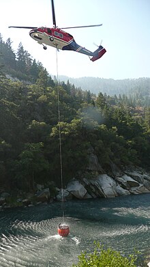 A helicopter dips its bucket into a river to drop water on a wildfire in California. Sikorsky S-70A-27 Picking Up Water.jpg