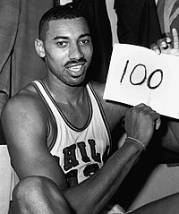 Wilt Chamberlain, an African American man, is shown sitting down in his Philadelphia Warriors jersey while holding up a piece of paper with the number 100 written on it. The photograph was taken directly after the game and is in black and white.