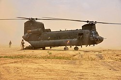 Colour photo of a dark green twin-rotored helicopter on the ground, with considerable amounts of dust in the air around it