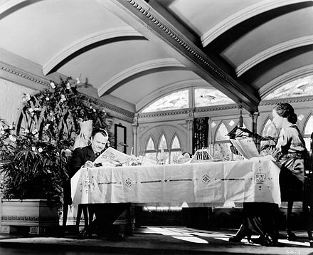 Two people seated at a large table in a garden room, reading newspapers