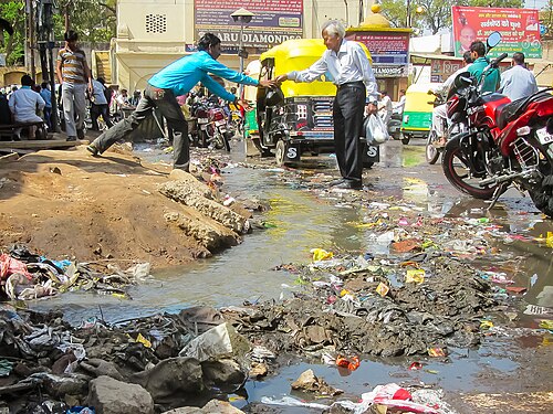 Man helping a senior cross a road in Mathura (India). The road is overflowing with sewage and garbage.