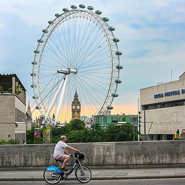 A man in London cycles on a Barclay-sponsored "Boris Bike" across Waterloo Bridge with the London Eye, Big Ben, and sunset in the background.