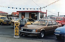 A used-car salesman may consider factors that signal the buyer's socioeconomic status, such as how the buyer is dressed and whether they sound educated, to decide what price to offer during price negotiations. Gary Lindbom, West Coast Car Court, Greymouth, 1987. CC147.jpg