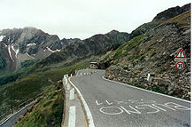 Uma estrada curva e ascendente, contra uma encosta rochosa.  Há inscrições em italiano na estrada, uma placa na beira da estrada e outros picos de montanhas visíveis ao fundo.