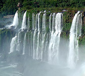 A section of the Iguazú Falls on the border of Argentina and Brazil. A bird in flight is visible on the left side of the image. Viewing platforms are visible in the upper left background.