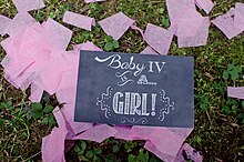 A black sign with white handwritten letters reads "Baby IV is a...girl!" The sign rests on grassy ground, and is surrounded by pieces of pink crepe paper confetti.