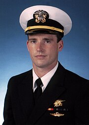 Color picture of Michael Murphy, a white male, wearing a military white dress hat and dark blue suit. There is a blue background behind him and he is wearing a gold Navy Seal Trident, 2 blue and green striped ribbons, 1 red and yellow striped ribbon and gold parachute insignia wings below the ribbons.