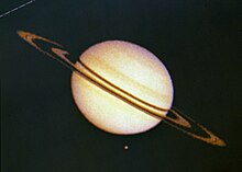 Saturno, no centro da imagem, totalmente iluminado, coloração bege. Ao seu redor, dois anéis marrons concêntricos, inclinados. Abaixo, um ponto laranja. Baixa definição da imagem.
