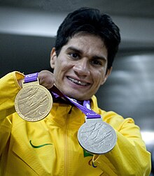 Yohansson Nascimento vestindo uniforme amarelo da delegação paralímpica brasileira mostra suas duas medalhas, uma de Ouro, outra de Prata, conquistadas na Paralimpíada de Londres-2012.