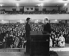 Two men stand facing each other on a platform as thousands of spectators on the ground floor and a balcony watch