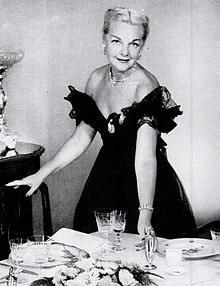 A blonde white woman posing in a low-cut black off-the-shoulder gown, behind a table set with dishes and glassware