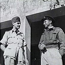 Deux hommes en uniforme militaire debout à l'extérieur d'un immeuble en train de se parler.  L'homme de gauche porte un uniforme de couleur plus claire à manches courtes.  L'homme de droite porte une veste sombre à manches longues.