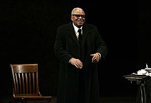 James Earl Jones standing on a stage with a black background with a wooden chair to his right and a table to his left while performing Driving Miss Daisy at the Theatre Royal in Sydney in March 2013