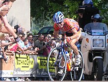 A cyclist in an orange jersey with purple trim, with many logos on it. He also wears sunglasses and a blue helmet, and sits crouched over his bicycle in an aerodynamic position. Spectators watch from behind barricades, and a cameraman follows on a motorbike.