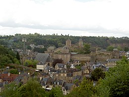 Photographie de Fougères. Dans un environnement boisé, on distingue des maisons au premier plan ainsi qu'une église et les remparts du château à l'arrière plan.