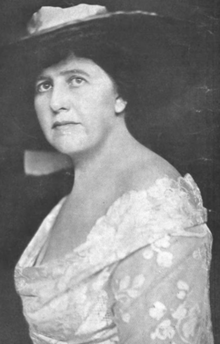 A middle-aged white woman, wearing a wide-brimmed hat and a white lace dress with a deep scoop, off-the-shoulder neckline