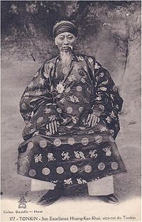 Aged man with cylindrical headdress, long flowing beard and moustache, a traditional Vietnamese tunic, darkly coloured with a light circular and Chinese character imprints, light coloured trousers, and dark shoes, sitting in a chair.