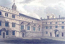 Stone buildings on two sides of a square, all topped with crenellations; from left, a three-storey building with a shell-hood above the door; a chapel with a bell turret and four gothic pointed windows, with the entrance porch under the furthest left window; this meets another three-storey building at the corner of the square, which has a large open archway in which two figures stand