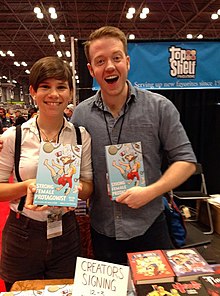 Seorang wanita kulit putih dan pria yang berdiri di sisi-by-side, menghadap kamera, dan tersenyum, mereka berdua memegang sebuah novel grafis berjudul "Protagonis Perempuan yang Kuat".