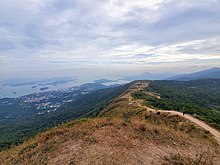 Hiking trail in foreground, winding down to a long, narrow plateau in the midground; small coastal town, inland sea and outlying islands in background