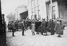 Photo noir et blanc. Un groupe de personnes avec bagages dans une rue, encadré par des soldats.