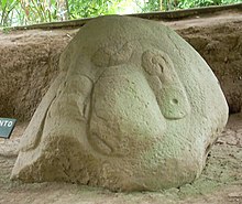 A large boulder set amongst the bare earth of an excavated area, with some vegetation visible in the background. The boulder has been carved into the form of an obese head with prominent puffed-out cheeks and swollen lips. The ears have large ear-spools hanging from them and the eyes are closed.