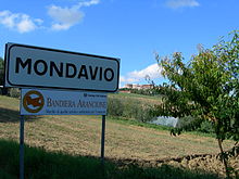 Il cartello stradale che segnala il comune di Mondavio. Sotto di esso, un cartello con il logo delle Bandiere Arancioni e la scritta: "Bandiera Arancione. Marchio di qualità turistico ambientale per l'entroterra". In alto a destra del cartello, il logo del Touring Club Italiano.