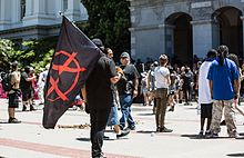 A crowd outside the California State Capitol after the riot. An anarchist flag is carried in the foreground. Debris lies on the plaza. Crowd outside California State Capitol after riot anarchist flag debris.jpg