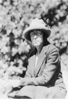 black and white of woman in glasses, felt hat and matching suit seated before trees.