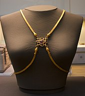Frontal view of a grey female bust with four gold bands of many fine links, two draped over the shoulders and two plunging from the breast bone sweeping below the breasts and going behind the back. The four converge between the breasts, where each band ends in a head which connects to a centrepiece, a purple gem surrounded by eight smaller sockets, four empty and four with red stones