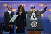 Barack Obama and Michelle Obama with Stewart celebrating the 75th anniversary of the USO in 2016 Jon Stewart tells jokes behind the president's lectern as President Obama and the First Lady look on during the comedy show in celebration of the 75th anniversary of the USO and the 5th anniversary of Joining Forces (26240098743).jpg